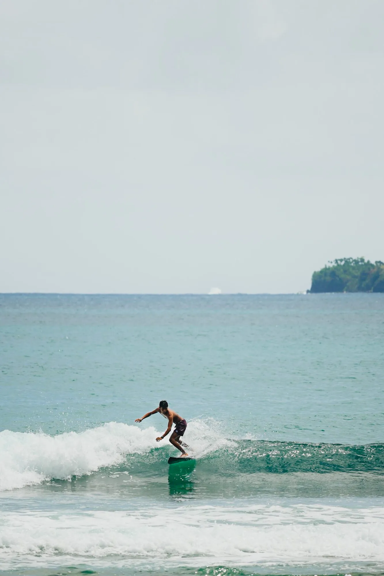A surfer riding a small green wave with arms out for balance and an island on the horizon